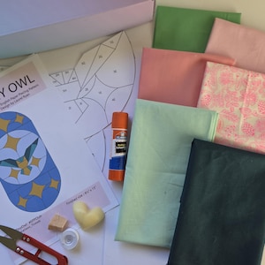 May include: A craft kit with fabric, scissors, and a glue stick. The kit includes a pattern for a "Tiny Owl" design, with a finished size of 8 x 15 inches. Fabric colors include green, pink, and blue. A heart-shaped wax melt is also included.