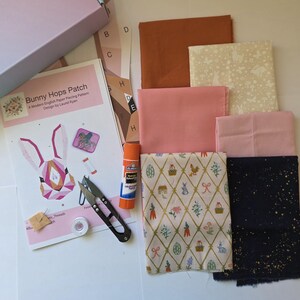 Könnte beinhalten: Ein Bastelset mit Stoffquadraten in verschiedenen Farben, darunter Rosa, Braun und gemusterte Stoffe. Das Set enthält ein Muster mit dem Titel "Bunny Hops Patch", eine Schere, Kleber und Faden. Für English Paper Piecing.