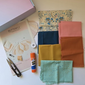 May include: A craft kit with fabric squares in various colors, including teal, mustard, and coral. The kit includes a butterfly pattern, scissors, glue stick, and thread. The box is light pink.