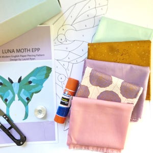 Könnte beinhalten: Ein Luna Moth EPP English Paper Piecing Schnittmuster von Laurel Ryan, ein Klebestift, eine Metallspule, ein schwarzer Nahttrenner und ein Stapel Stoffquadrate in Minze, Senfgelb, Lavendel, Blumenmuster und Hellrosa, mit Papierschablone.