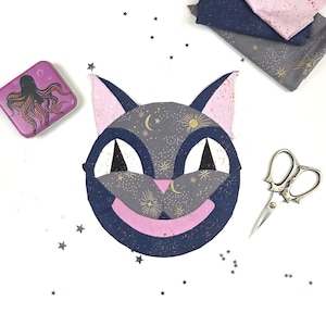 May include: A whimsical cat face design with a navy blue outline, grey face, and pink accents. The cat's face is decorated with gold celestial patterns. A purple tin with an octopus design, fabric scraps, and scissors are also in the image.