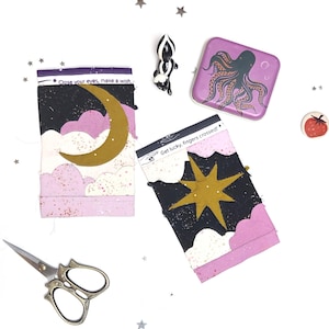 May include: A collection of decorative items on a white background. Features two fabric pieces with celestial designs and text, a skunk figurine, a purple octopus pin, a strawberry button, and decorative scissors. The fabric pieces have gold accents and the text "Close your eyes, make a wish" and "Get lucky, fingers crossed!"