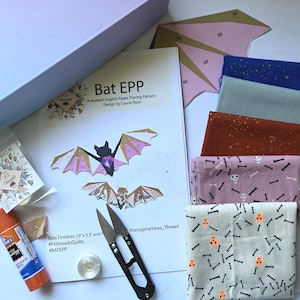 May include: A craft kit for English paper piecing featuring a Bat EPP pattern by Laurel Ryan, fabric swatches in blue, grey, rust, and skull prints, a glue stick, metal snips, paper templates, and a small container of thread balm.