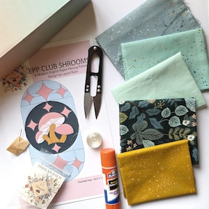 May include: A craft kit featuring a paper pattern titled EPP CLUB SHROOMS by Laurel Ryan, four folded fabric swatches in blue, mint, dark green floral, and mustard yellow, black thread snips, a glue stick, a metal bobbin, and thread balm.