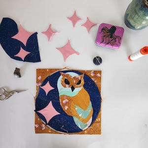 May include: A close-up of a fabric owl applique with pink stars on a blue background. The owl is surrounded by a brown fabric border with floral designs. There are several pink fabric stars scattered around the owl applique.
