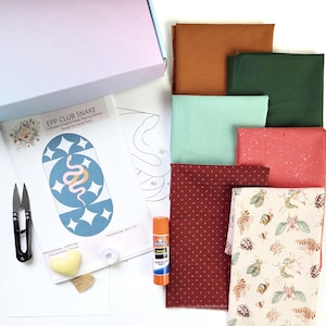 May include: A craft kit with fabric squares in various colors, a pattern with a snake design, scissors, glue, and other supplies. The pattern is titled "EPP CLUB SNAKE" and includes the text "A Modern English Paper Piecing Pattern".