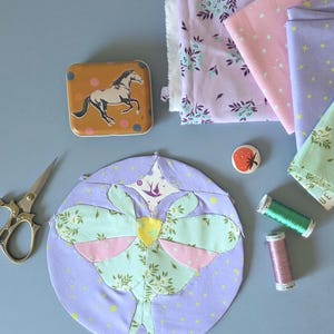 May include: A craft scene with a circular fabric piece featuring a moth design in pastel colors. A horse tin, fabric scraps, scissors, thread spools, and a strawberry pin are arranged on a blue surface. The fabrics have floral and star patterns.