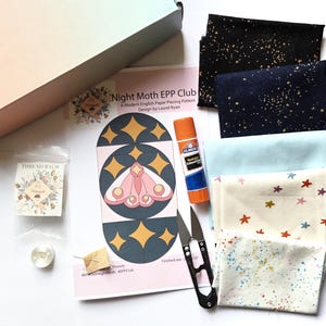 May include: A craft kit containing a Night Moth EPP Club pattern sheet, an orange Elmer's glue stick, black metal thread snips, a small clear bag of thread balm, a white button, and several folded fabric squares in black, navy, light blue, and cream.