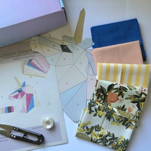 Op de afbeelding: Een knutselpakket met een papieren sjabloon van een geometrische eenhoornkop, een stapel gevouwen stoffen vierkanten in blauw, perzik, gele strepen en bloemmotieven, een metalen spoeltje en een metalen schaar, inclusief een Unicorn Dream instructieblad.