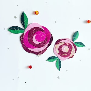 May include: Two paper roses with pink and burgundy petals and gold accents. The roses have green leaves.