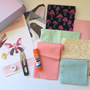 Könnte beinhalten: Ein Bastelset mit Stoff, Papiermustern, Schere und Kleber. Der Stoff umfasst einen schwarzen Pilzdruck, rosa, mintgrüne und sternförmige Stoffe. Das Set enthält auch ein Papiermuster für ein Waschbären-Design.