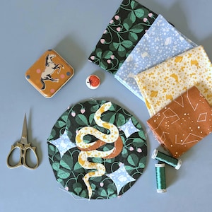 May include: A selection of craft materials: patterned fabric squares, a circular fabric piece with a snake applique, a small tin with a horse illustration, scissors, thread spools, and a strawberry button. The fabrics display botanical and celestial patterns.