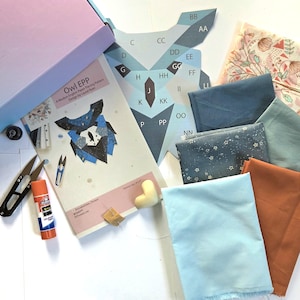 Könnte beinhalten: Eine Auswahl an Quiltzubehör, darunter Stoff, Schere, Kleber und ein Nähmuster für eine Eule. Die Stoffstücke sind in verschiedenen Farben gehalten, darunter Blau, Orange und Hellblau. Das Muster trägt den Titel "Owl EPP".