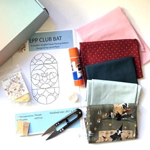 May include: A quilting kit featuring an EPP Club Bat pattern sheet, a glue stick, thread balm, small scissors, and five folded fabric pieces in pink, dark red with stars, dark green, light blue, and a badger-themed print.