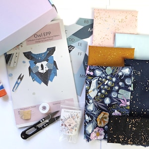 Könnte beinhalten: Ein Bastelset mit einer Owl EPP-Anleitung, Stoffstücken in Rosa, Hellblau, Senfgelb und Dunkelblau mit Blumen- und Sprenkelmustern, einem Klebestift, Garnbalsam, einer kleinen Spule mit weißem Garn und einer Metallschere.