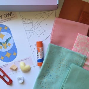 Könnte beinhalten: Ein Bastelset mit Stoffquadraten, einem Papiermuster und einem Klebestift. Das Set enthält ein gedrucktes Eulen-Design, eine Schere und eine herzförmige Kerze. Der Stoff ist in den Farbtönen Rosa, Braun und Türkis gehalten.
