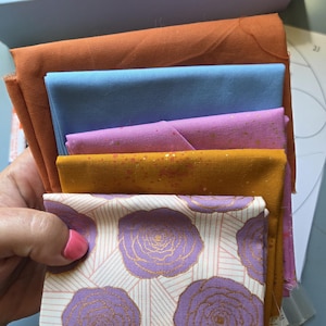May include: A stack of five folded fabric squares featuring a variety of colors and patterns. The collection includes solid burnt orange, light blue, and mustard yellow with gold speckles, a pink fabric with gold speckles, and a white floral print.