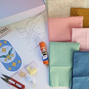 Könnte beinhalten: Ein Bastelset mit Stoff, Schere, Kleber und einer Papierschablonen-Vorlage für ein Eulen-Design. Das Set enthält Stoffe in verschiedenen Farben, darunter Rosa, Grün und Blau. Die fertige Größe der Eule beträgt 20,3 cm x 38,1 cm.