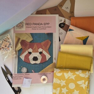 May include: A Red Panda EPP English paper piecing pattern by Laurel Ryan, featuring a red panda illustration, alongside fabric swatches in orange, yellow, and star patterns, a small packet of thread balm, metal snips, and paper templates.