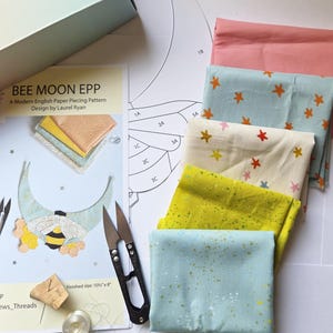 May include: A Bee Moon EPP English paper piecing pattern by Laurel Ryan, featuring a printed guide, a stack of five folded fabric pieces in pink, light blue, cream, and yellow, metal thread snips, a needle in a cork, and a metal bobbin.