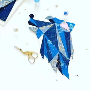 May include: A blue and gray fabric wolf head, made of patchwork fabric pieces, is displayed on a white surface. The wolf head is a geometric design with a pink nose. A pair of gold scissors is also in the image.