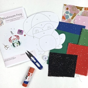 May include: A paper piecing pattern named "Mushroom Frog" featuring a frog design, alongside fabric squares in diverse colours and patterns. Includes scissors, glue stick, and thread. The finished size is 22.86 cm x 22.86 cm.