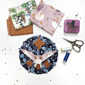 May include: An overhead shot of quilting supplies. Includes fabric squares with floral, swan, and constellation patterns. A partially sewn quilt block, a purple octopus tin, scissors, and spools of thread are also present. The color palette is a mix of blues, browns, pinks, and greens.