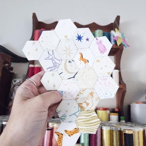 May include: A handmade, hexagonal patchwork piece featuring various whimsical designs. The fabric pieces are white with illustrations of a fox, horse, stars, and other celestial and animal motifs. The piece is held in a hand, with spools of thread in the background.