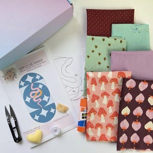 Puede incluir: Una caja abierta revela un kit de proyecto de costura con paquetes de tela, un patrón y herramientas. El patrón presenta un diseño azul y blanco con una ilustración de serpiente. Los paquetes de tela incluyen patrones de estrellas, setas y fresas. También se ven tijeras, pegamento e hilo.