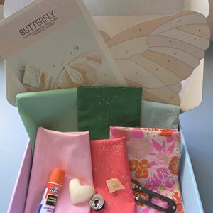 Op de afbeelding: Een knutseldoos gevuld met stoffen, een lijmstift en een hartvormig item. De doos bevat een patroon met het label "BUTTERFLY" en verschillende stoffen in roze, groene en bloemmotieven. Een kleine schaar en een spoel zijn ook inbegrepen.