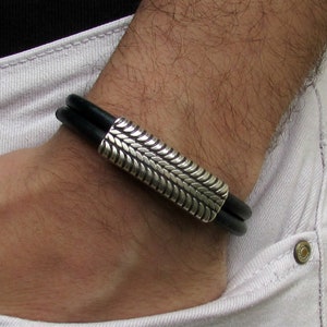 Könnte beinhalten: Ein schwarzes Lederarmband mit einem silbernen, strukturierten, rechteckigen Verschluss.