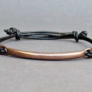 Op de afbeelding: Een eenvoudige armband met een koperen staaf en een zwart leren koord. Het koord is aan beide uiteinden met een knoop vastgemaakt.