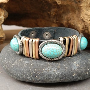 May include: A black leather bracelet with turquoise stones and metal accents. The bracelet has a snap closure and is decorated with copper and silver colored metal bands.