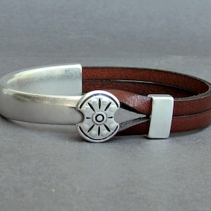 Puede incluir: Una pulsera de cuero marrón con cierre plateado y centro decorativo. La pulsera presenta dos correas de cuero y un elemento decorativo plateado con un diseño circular. La pulsera mide aproximadamente 20 cm de largo.