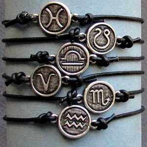 May include: A set of twelve black leather bracelets with silver charms featuring the astrological signs. The charms are arranged in a circle, with each sign facing outward.