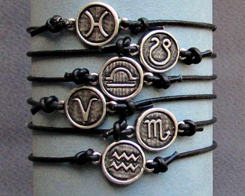 May include: A set of twelve black leather bracelets with silver charms featuring the astrological signs. The charms are arranged in a circle, with each sign facing outward.