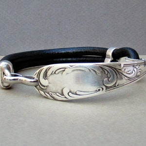May include: A silver bracelet with an ornate design, attached to a black leather band.