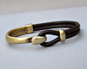 May include: A brown leather bracelet with a gold metal clasp. The clasp is a simple loop design.