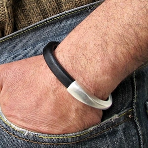May include: A black leather bracelet with a silver metal clasp. The bracelet is worn on a person's wrist.