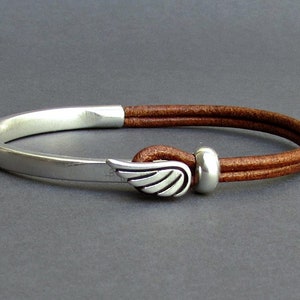 May include: A brown leather bracelet with a silver wing charm and a silver bead. The bracelet is open and has a silver metal band.
