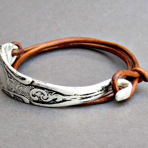 May include: A silver spoon bracelet with an ornate design, attached to a brown leather strap.