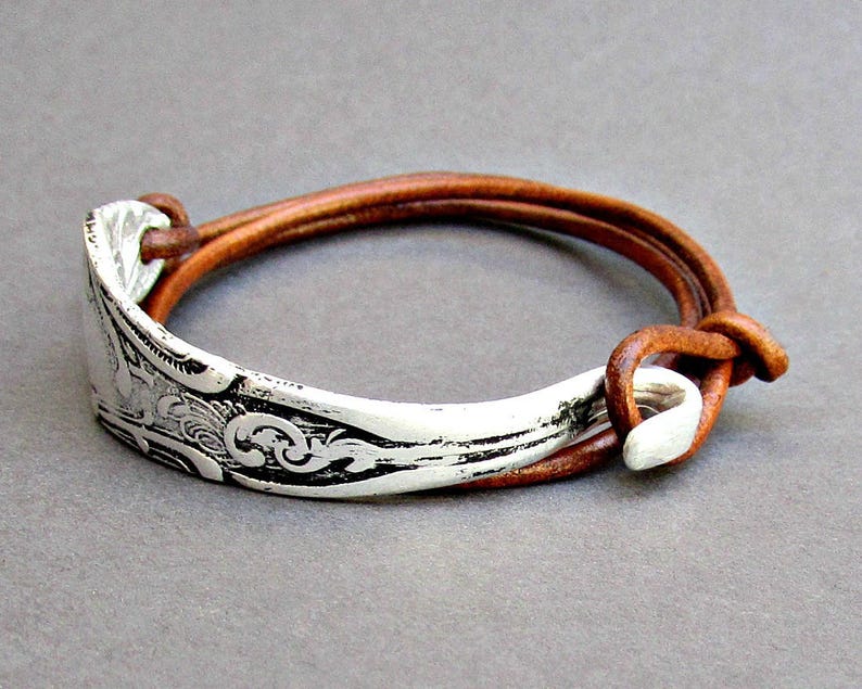 May include: A silver spoon bracelet with an ornate design, attached to a brown leather strap.
