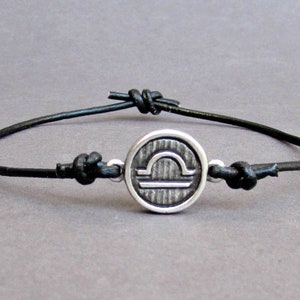 May include: A black leather bracelet with a silver charm featuring the Libra zodiac symbol.