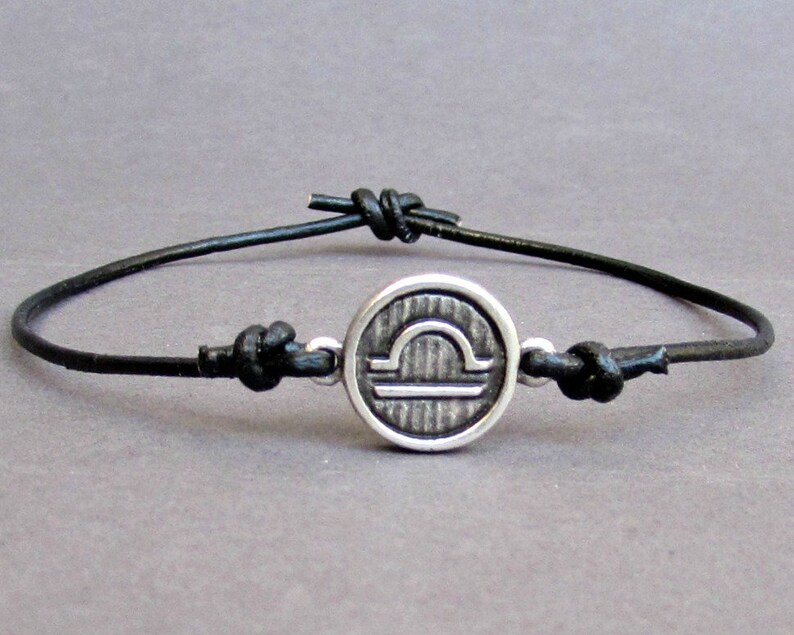May include: A black leather bracelet with a silver charm featuring the Libra zodiac symbol.