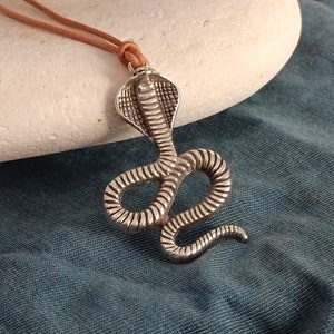 Puede incluir: Un collar con un colgante de cobra de plata con un cordón de cuero marrón. El colgante está detallado con escamas texturizadas y una cabeza de cobra en relieve.