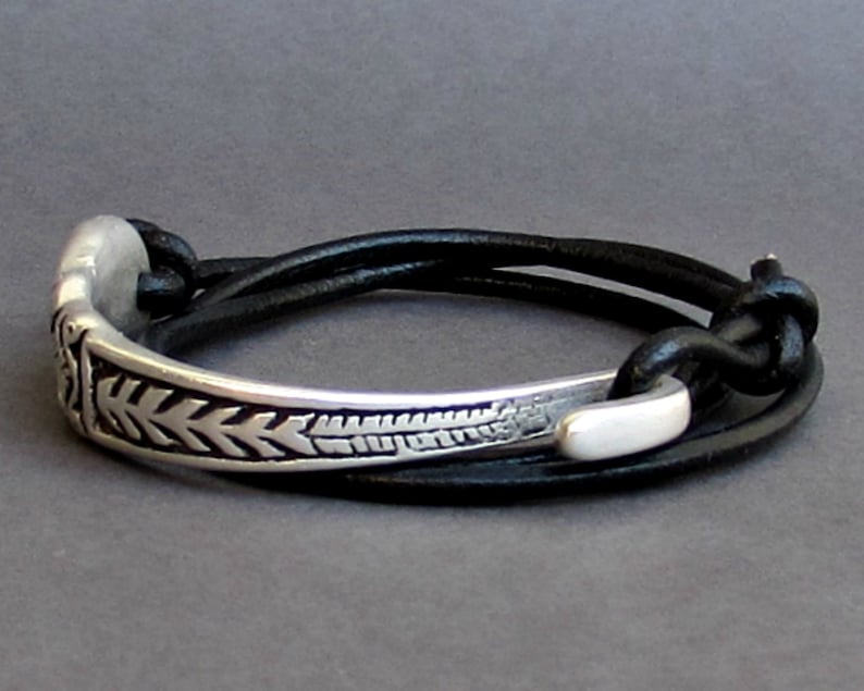 May include: A silver bracelet with a decorative design and a black leather strap. The strap is adjustable and can be worn in multiple ways.