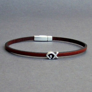 Puede incluir: Un brazalete de cuero marrón con un dije de plata en forma de pez. El brazalete tiene un cierre magnético.