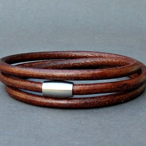 May include: A brown leather bracelet with a silver metal accent. The bracelet is made of three strands of leather that are woven together.