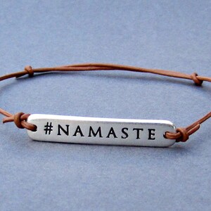 Puede incluir: Un brazalete de cuero marr&oacute;n con un dije de plata que dice "#NAMASTE".