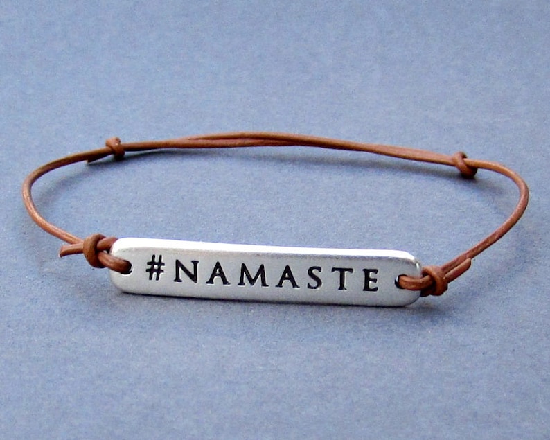 Puede incluir: Un brazalete de cuero marr&oacute;n con un dije de plata que dice "#NAMASTE".